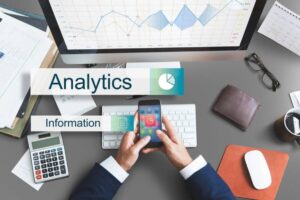 A professional analyzing data and graphs on a smartphone and computer surrounded by business analytic charts and office supplies.