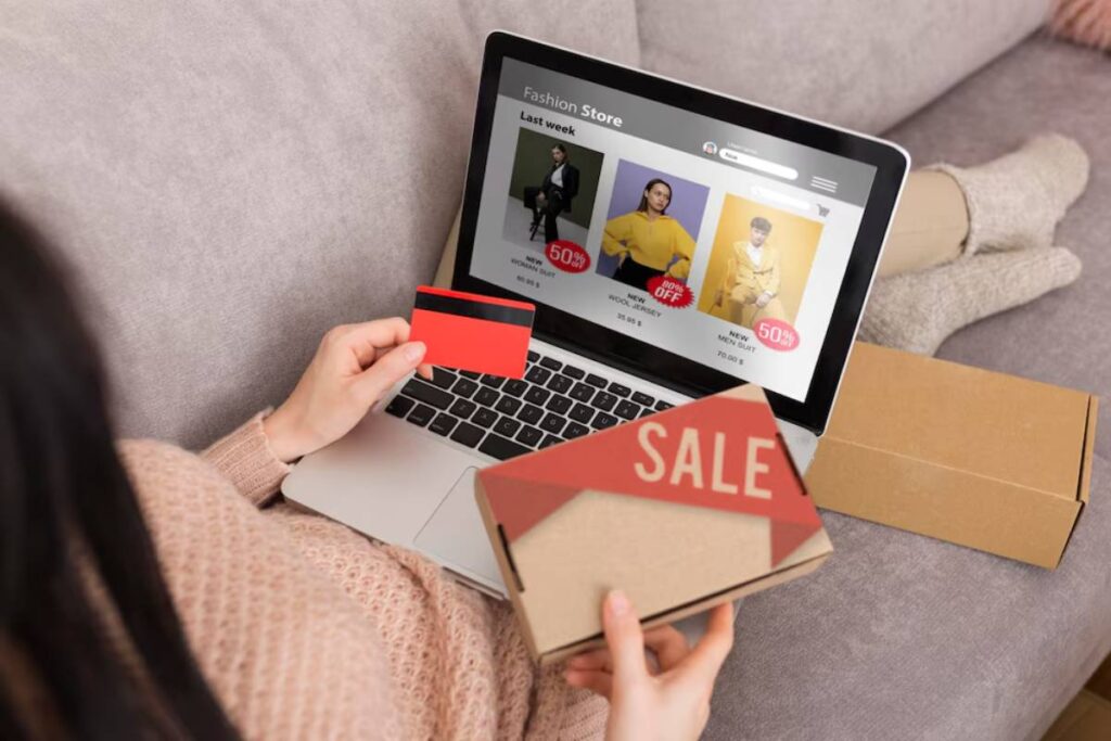 Woman shopping online on a laptop, holding a credit card and a sale flyer, with packages nearby.