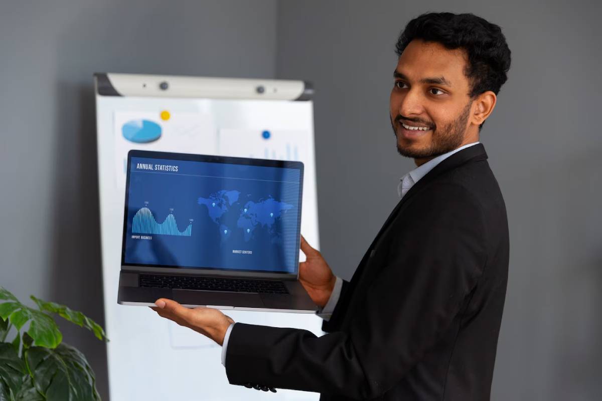 A businessman smiling, holding a laptop displaying graphs and world map statistics in an office setting.