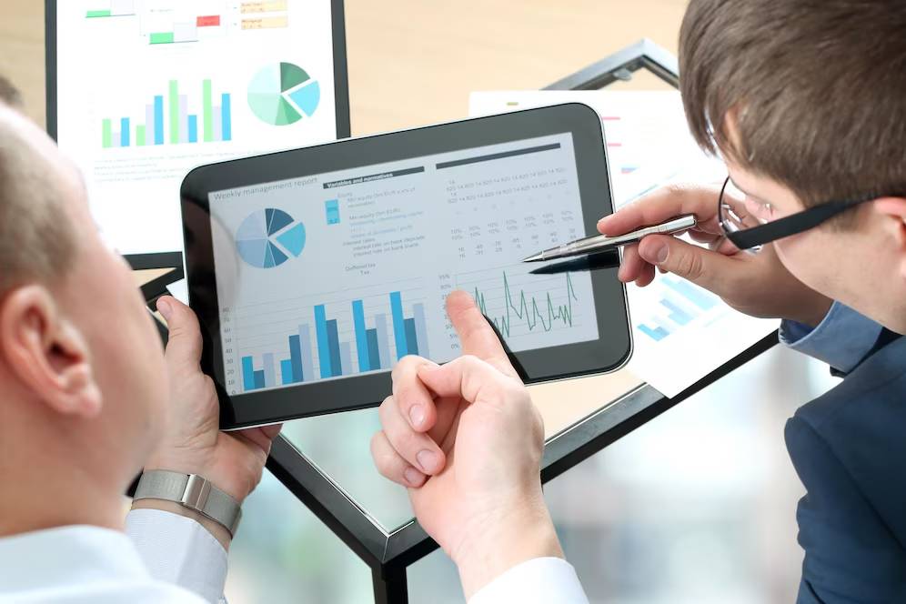Two professionals analyzing financial data charts on a digital tablet and screen in an office setting.
