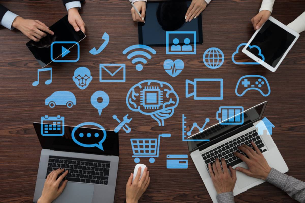 Overhead view of four people using digital devices surrounded by various technology and communication icons on a wooden table.