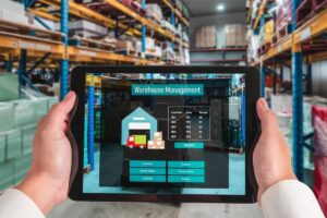 Person holding a tablet displaying a warehouse management system with a warehouse in the background.