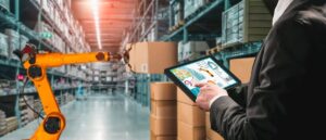 A person using a tablet to control a robotic arm in a warehouse with boxes.