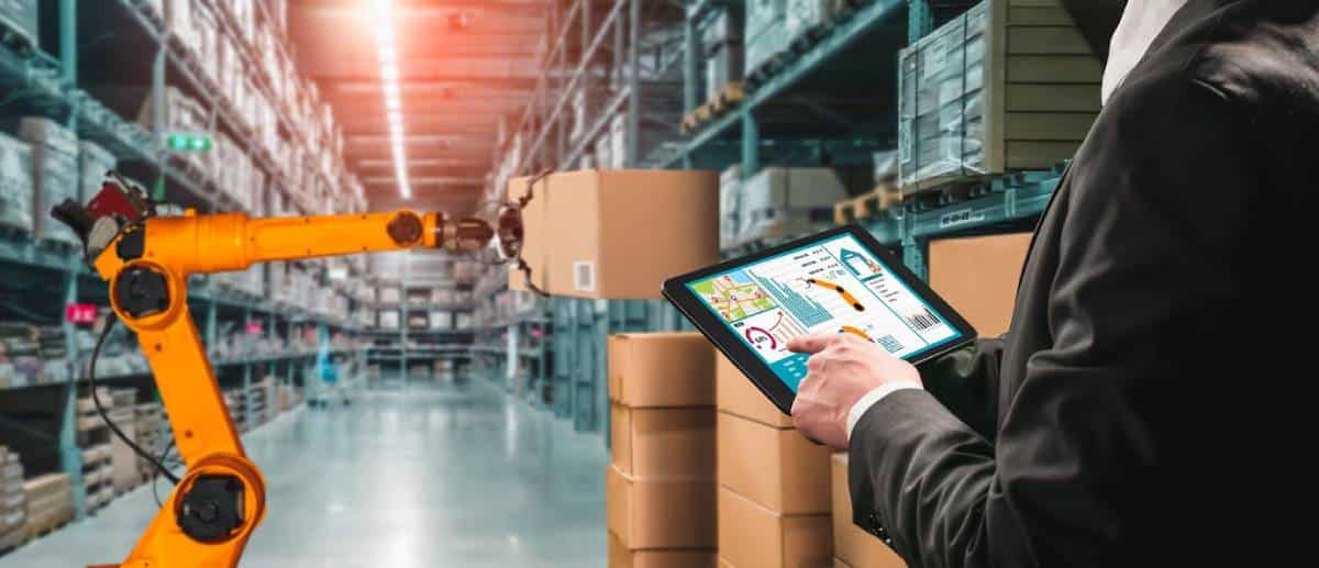 A person using a tablet to control a robotic arm in a warehouse with boxes.