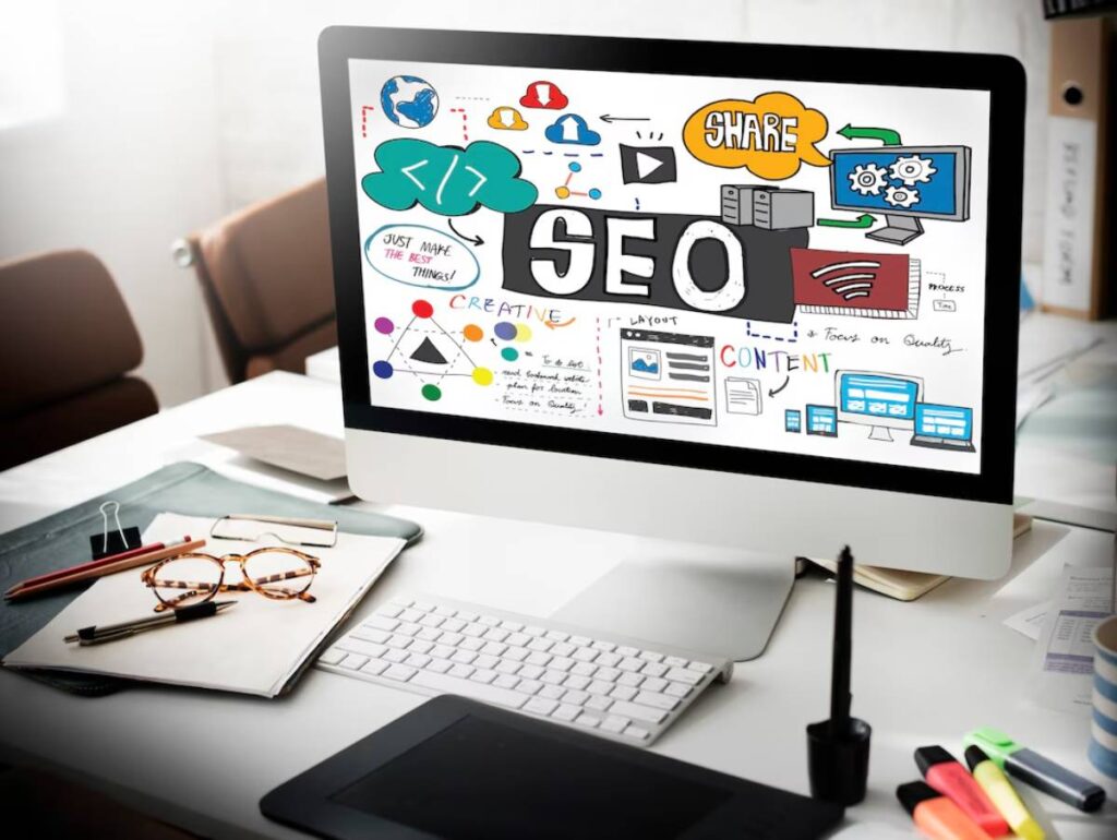Desktop computer showing colorful SEO and digital marketing concepts on screen, surrounded by office supplies on a desk.