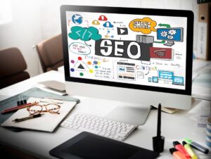 Desktop computer showing colorful SEO and digital marketing concepts on screen, surrounded by office supplies on a desk.
