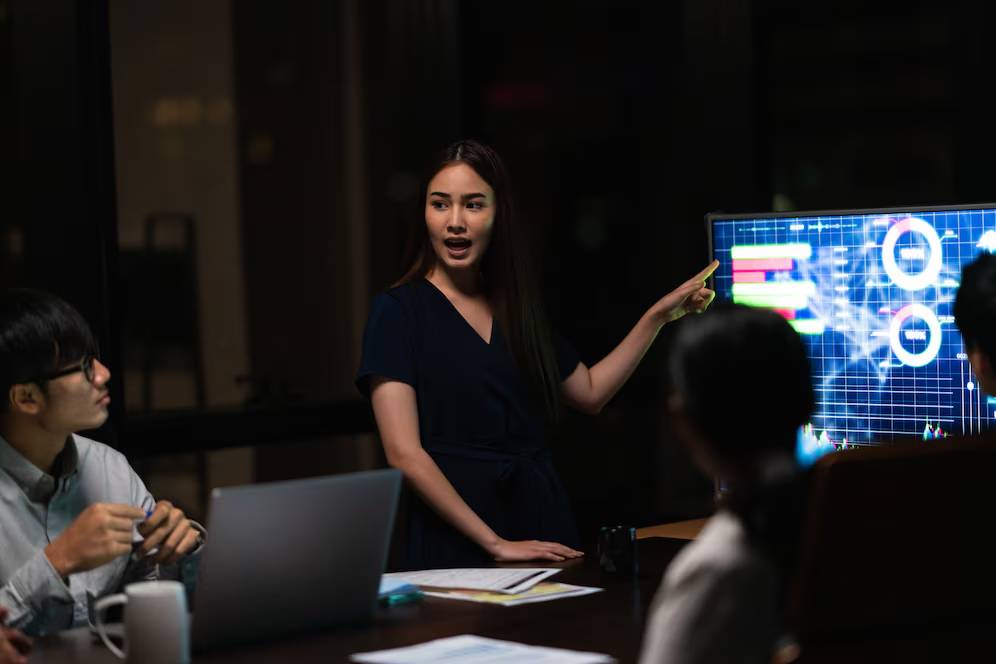 A woman presents data on a screen during a meeting in a dimly lit office.