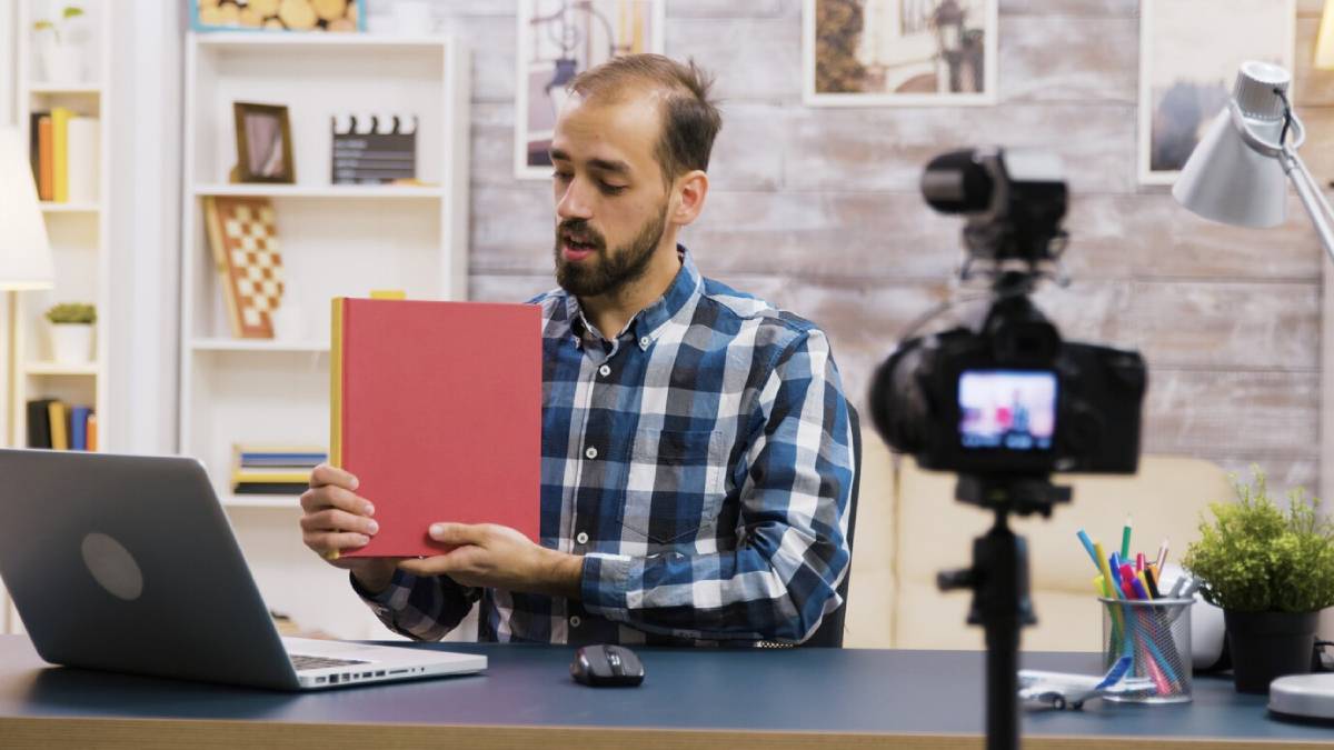 Man in plaid shirt showing a book to a camera in a home office setting.